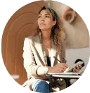 A woman in a light-colored blazer holds a pen over a notebook, looking thoughtfully off-camera.