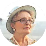 A circular headshot of an older woman wearing glasses, a hat, and a pearl earring.