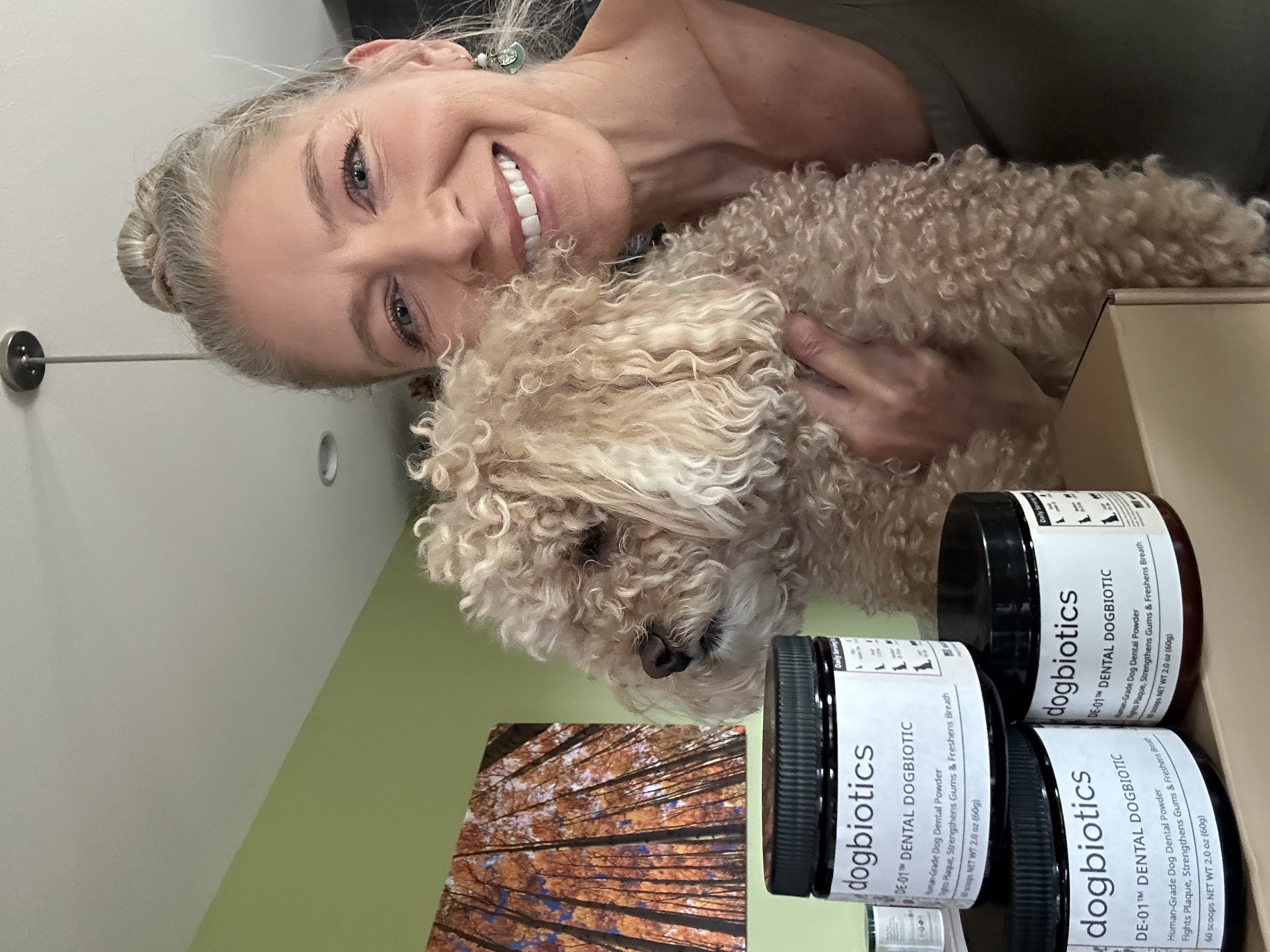 Smiling woman holds a fluffy dog beside jars labeled 'dogbiotics' in a room with green walls.