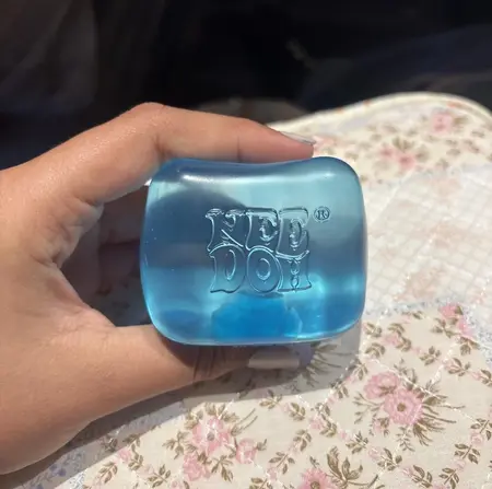 A person's hand holding a blue, cube-shaped, translucent NeeDoh toy over a floral-patterned surface.