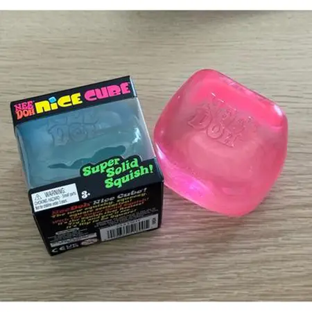 A pink, translucent Nee Doh Nice Cube squishy toy next to its black packaging on a wooden surface.