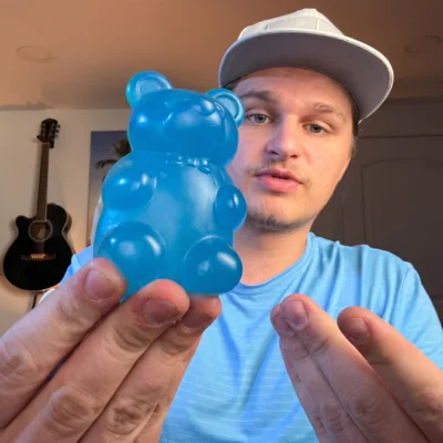A man in a backwards baseball cap holds up a large, translucent blue gummy bear.