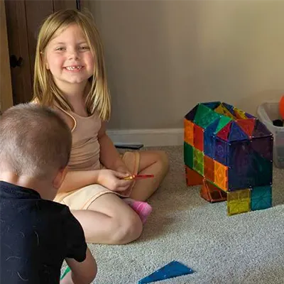 Two children building with colorful magnetic tiles on a carpeted floor.