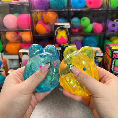 Two hands hold blue and yellow translucent, glittery bear-shaped toys in front of a toy display.