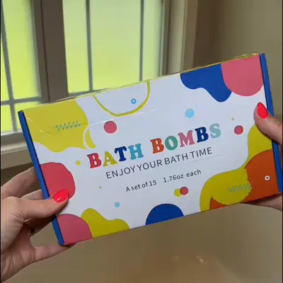 A person's hands with bright pink nail polish holding a colorful box of bath bombs.