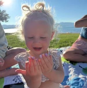 Child looking at painted fingernails while sitting on a blanket by a sunny lakeside.