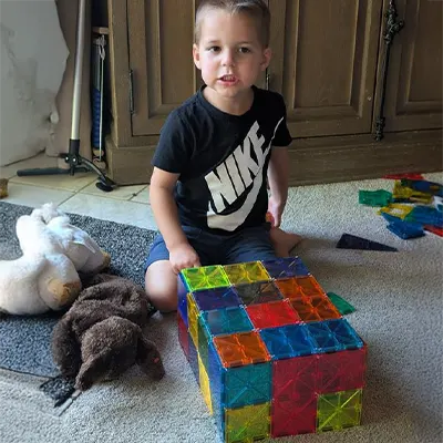 Child sits on carpet, playing with colorful magnetic tiles cube next to stuffed animals.