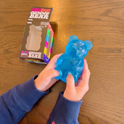 A person holds a large, blue, squishy gummy bear toy with its packaging on the table.