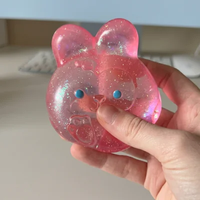A hand holds a translucent pink, glittery toy shaped like a bunny's head with blue eyes.