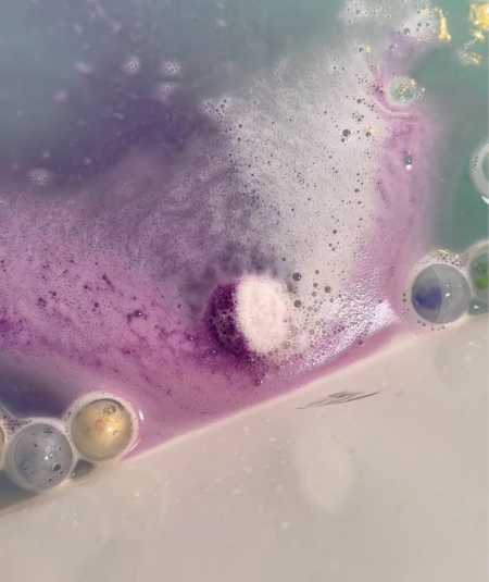 A purple and white bath bomb fizzing in a bathtub, creating colorful foam and bubbles.