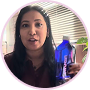 A woman with long dark hair holds a blue bottle while speaking to the camera.