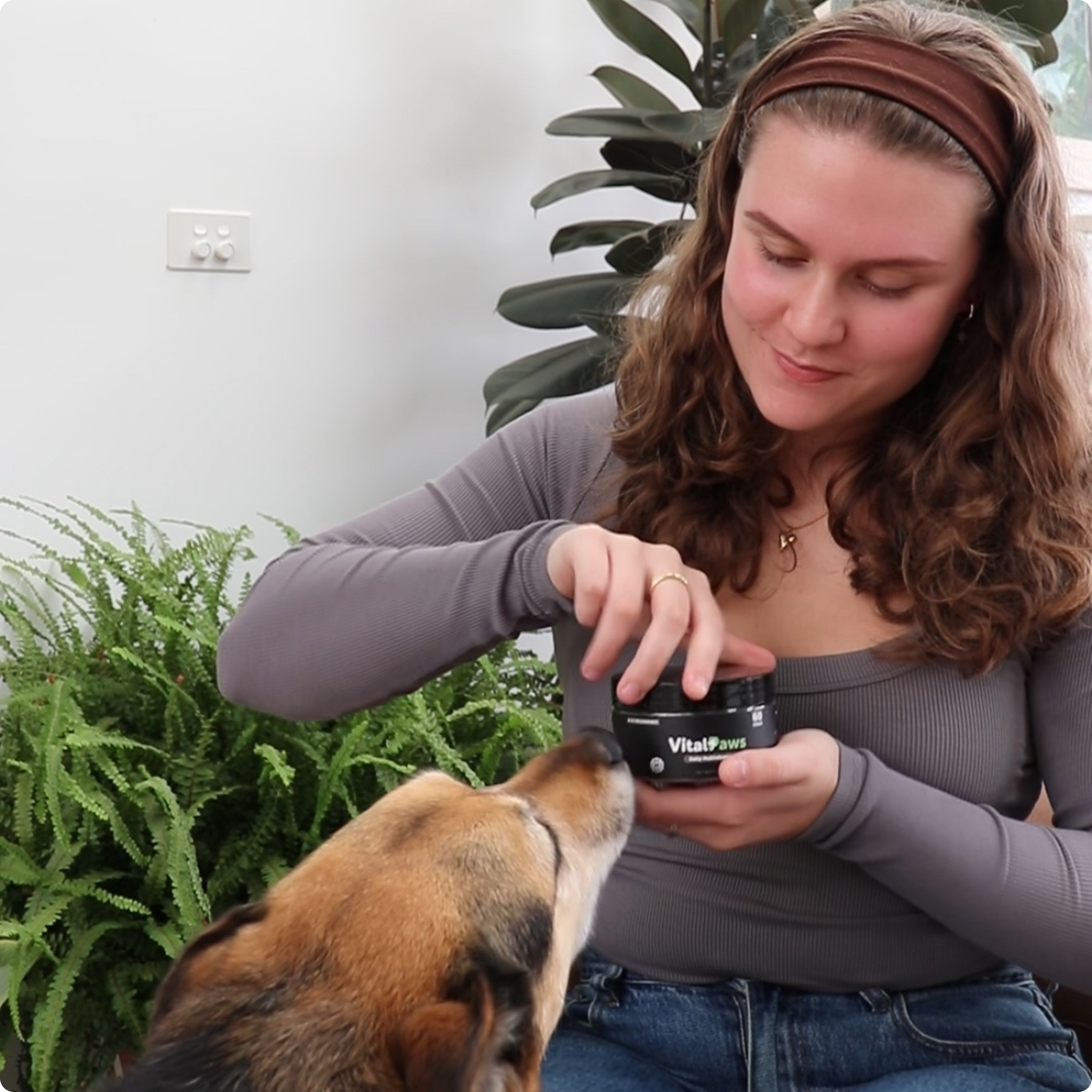Person opening a jar labeled 'Vital Paws' with a dog nearby.