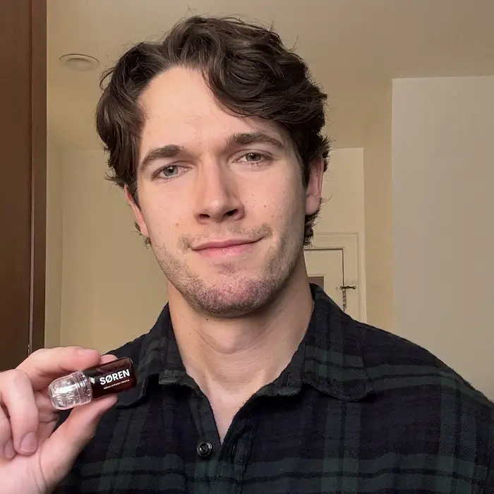 A man in a plaid shirt smiles while holding a small vial labeled SØREN.