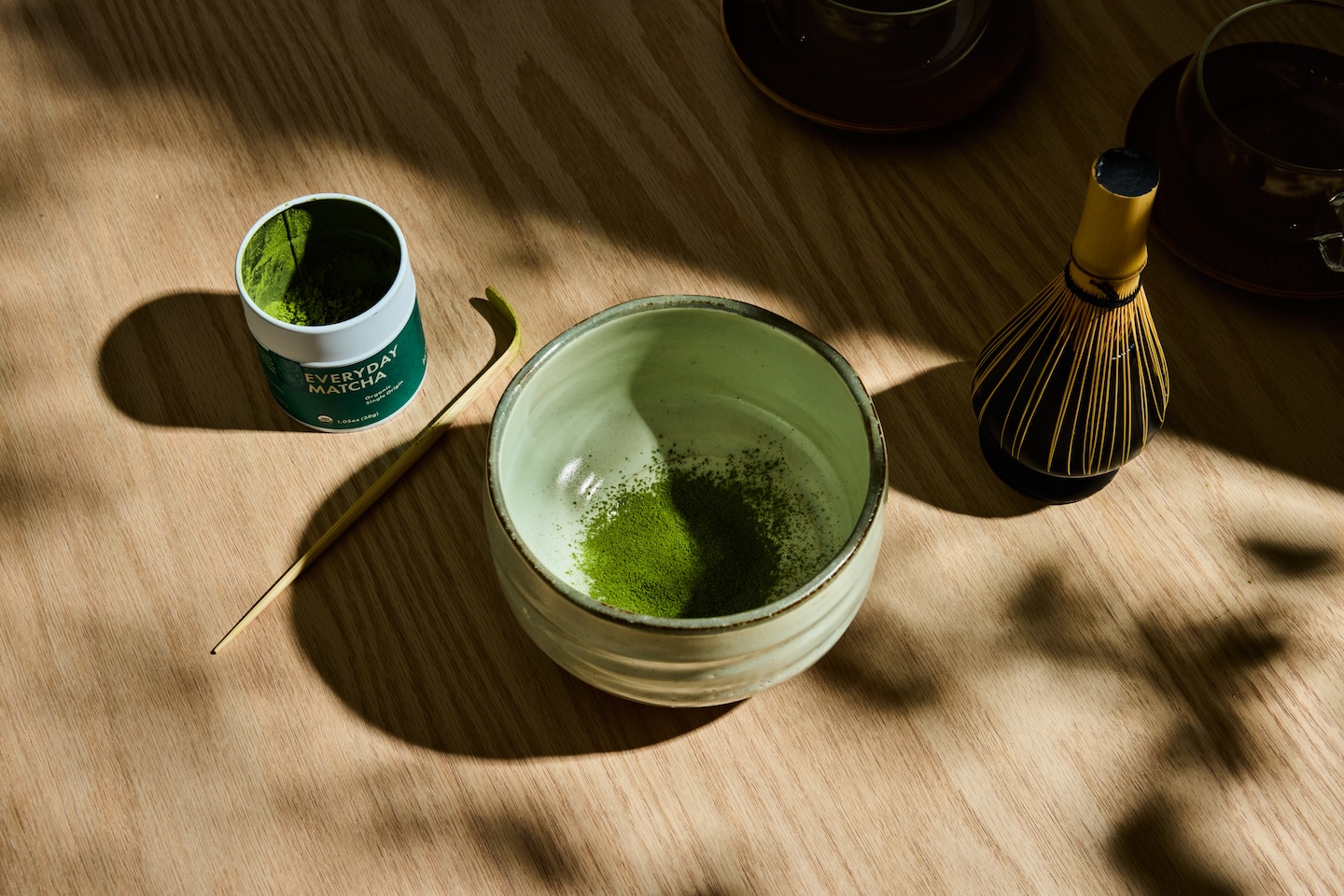 Matcha tea setup with bowl, whisk, and open matcha container on wooden table.