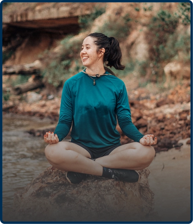 Person meditating outdoors on a rock by a riverbank.