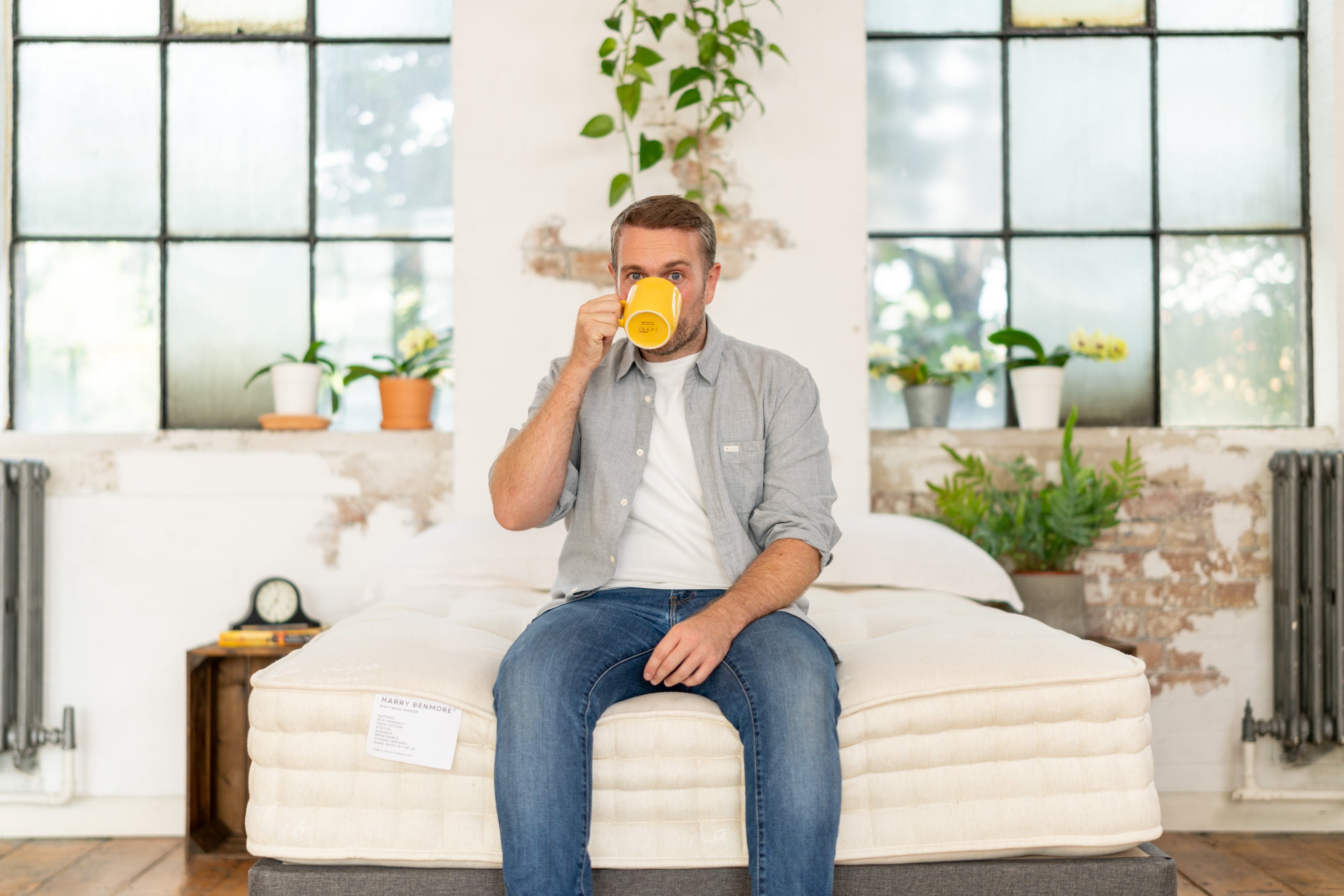Man sitting on bed holding a yellow mug indoors with plants.