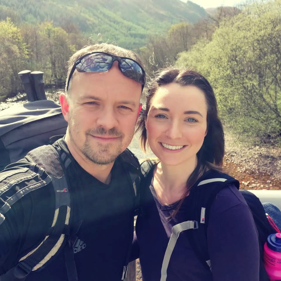 Two people with backpacks smiling outdoors in a forested area.