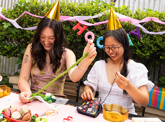 Two people wearing party hats crafting with decorations and macarons on the table.