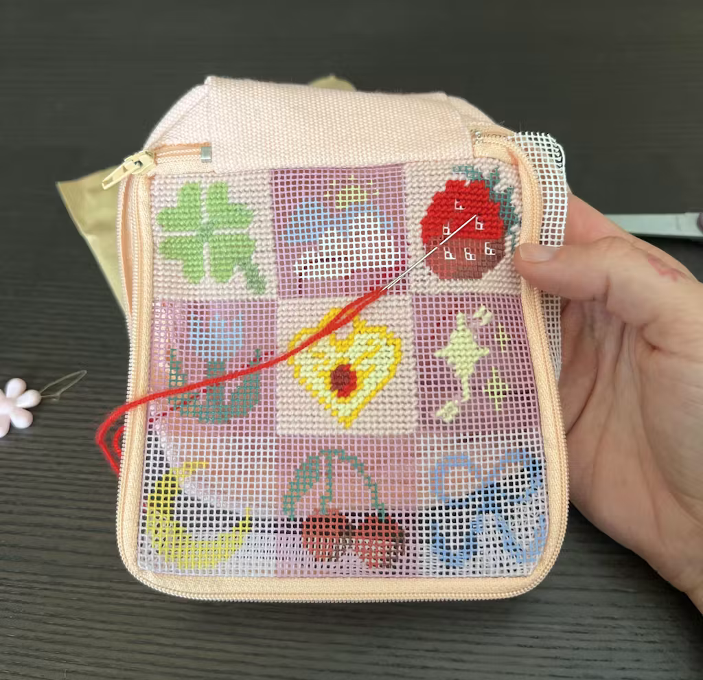 Cross-stitch embroidery in progress on a small, pastel pink bag with floral and fruit designs.