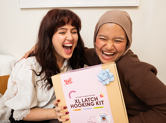 Two smiling people holding an XL latch hooking kit.