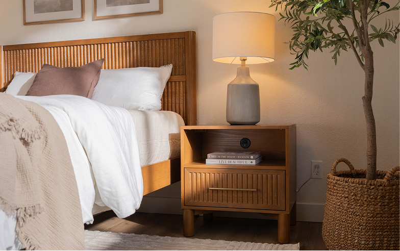 A warm bedroom with wooden nightstand and matching bed. The nightstand has books on its shelf, and a lamp on top, casting warm lighting onto the wall behind it.