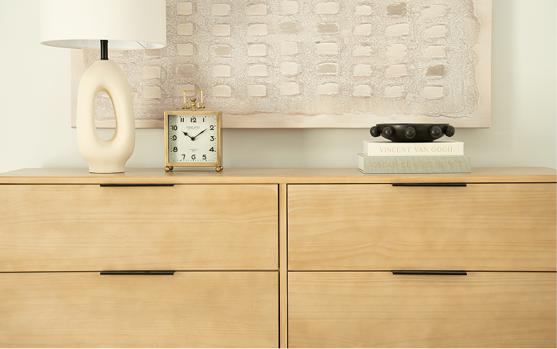A close-up of a light wood dresser. On top of the dresser is a lamp, clock, books, and a bowl. On the wall behind the dresser is a framed painting.