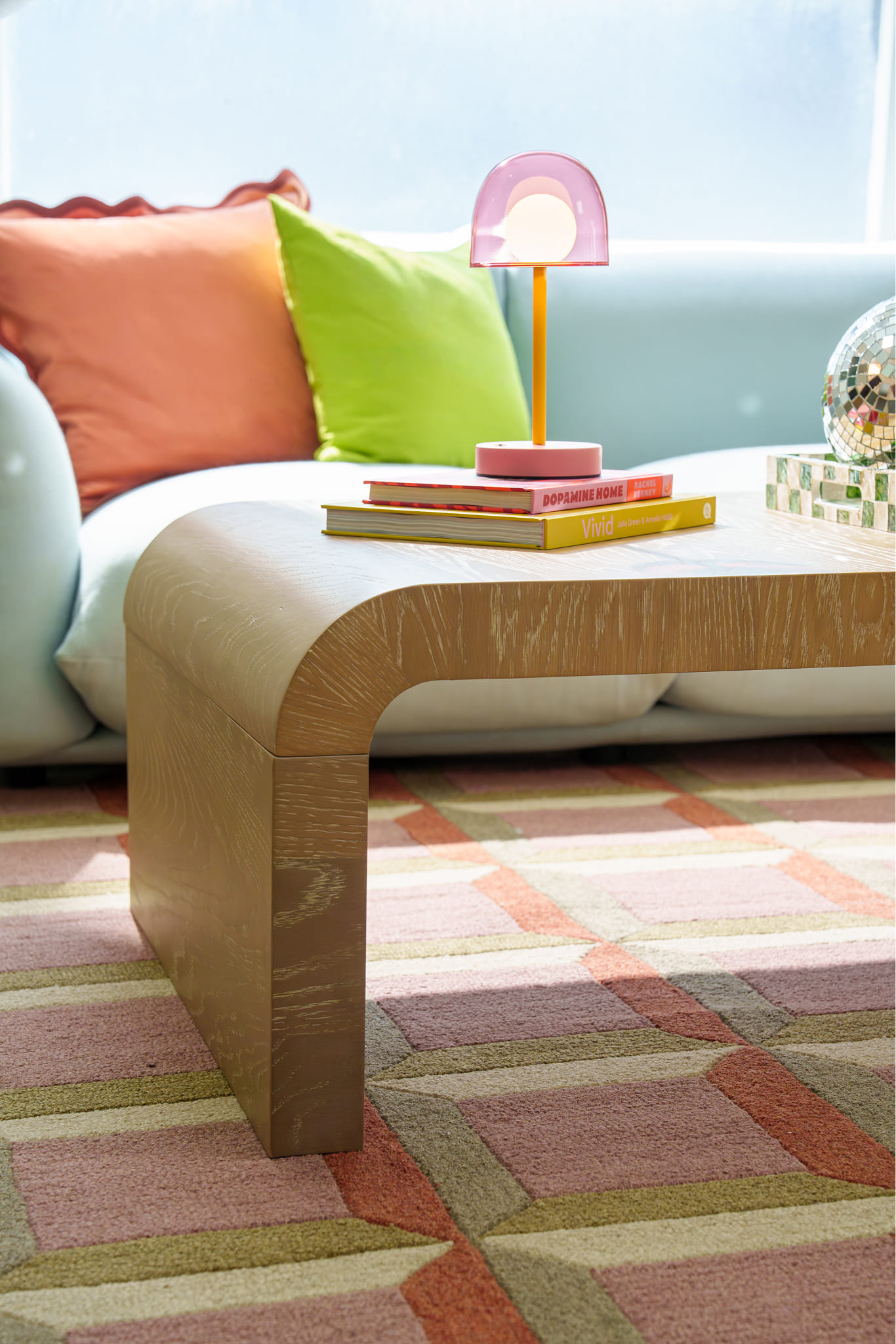 A light wood waterfall coffee table with colorful books sits on a geometric patterned rug.