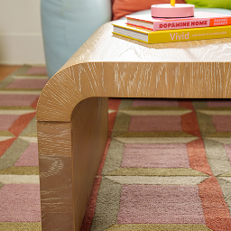 A light wood waterfall coffee table with colorful books sits on a geometric patterned rug.