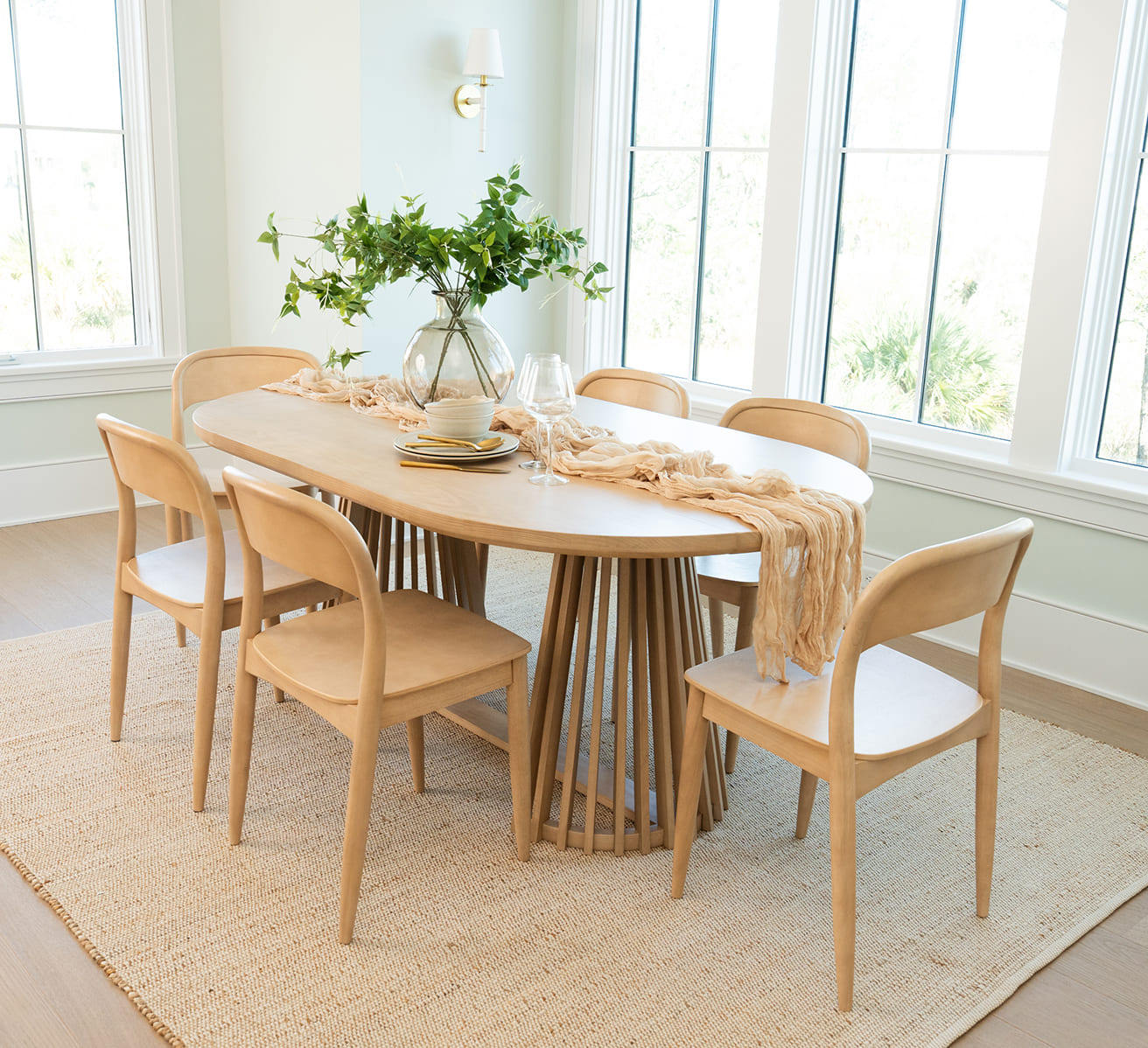 Light wood dining table with matching chairs, decorated with table runner, dishes, wine glasses, and vase with greenery.
