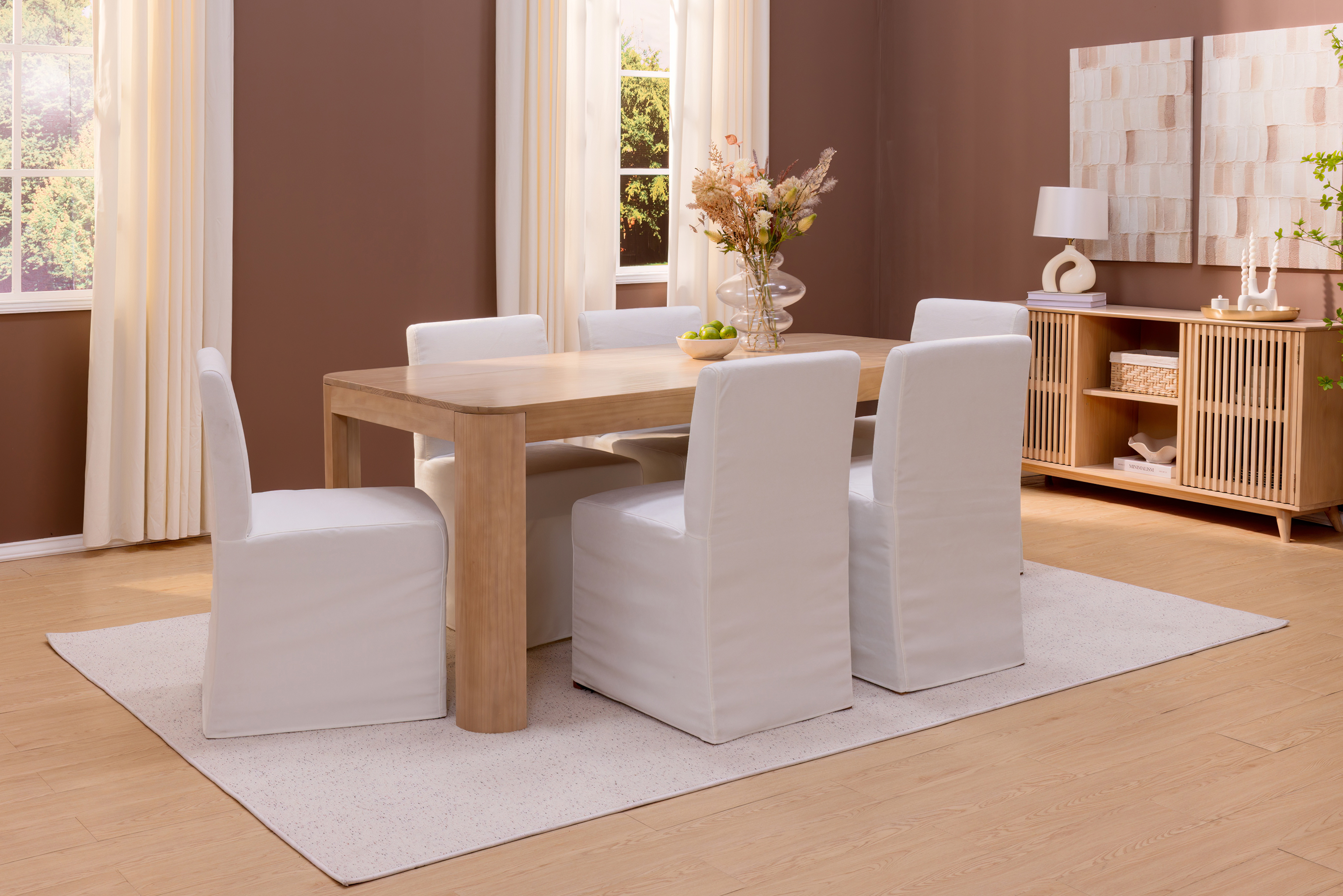 A warm dining room with light wood dining table, white cloth covered dining chairs, and matching white rug.