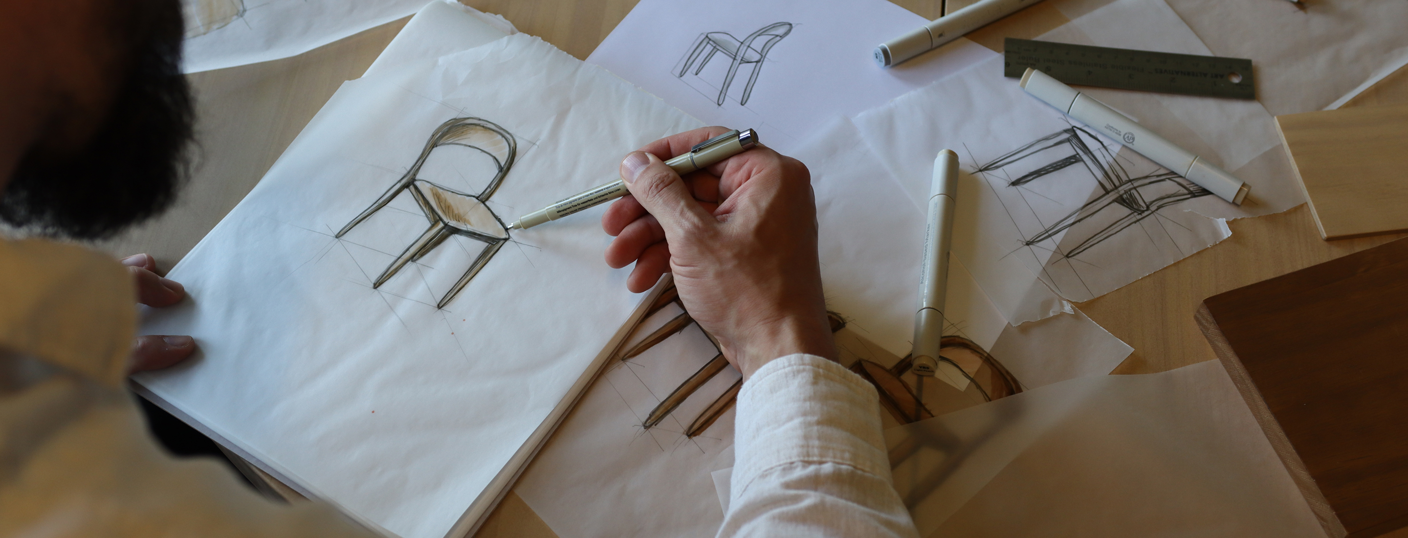 A bearded man sketching a wooden dining chair. Around the sketch are various other sketches of the same chair in different angles, as well as some pens.