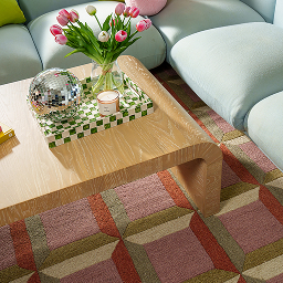 A light wood waterfall coffee table with colorful books sits on a geometric patterned rug ariel view.