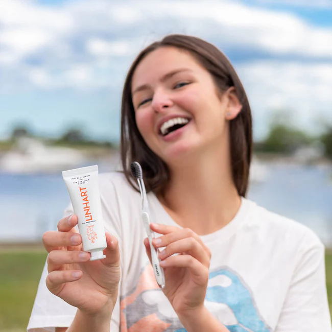 Person smiling with a toothbrush and toothpaste outdoors.