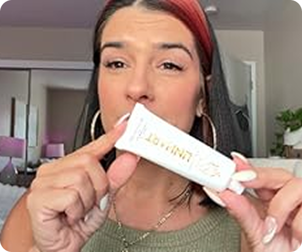 Person holding a tube of cream, pointing to it in a room with a mirror and plants.