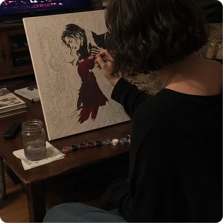 A person with short, dark hair works on a paint-by-numbers canvas of a couple dancing.