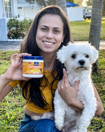 Woman holding a small white dog and a container, sitting on grass.