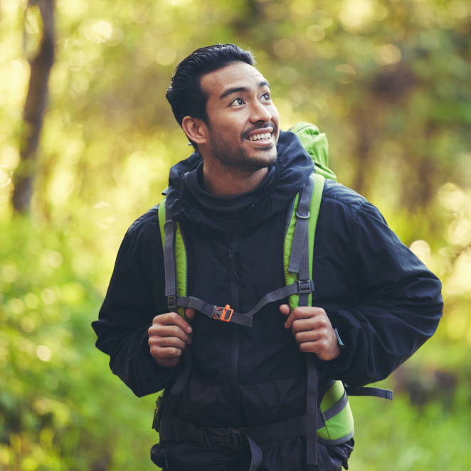 Man hiking in a forest with a backpack, looking up and smiling.