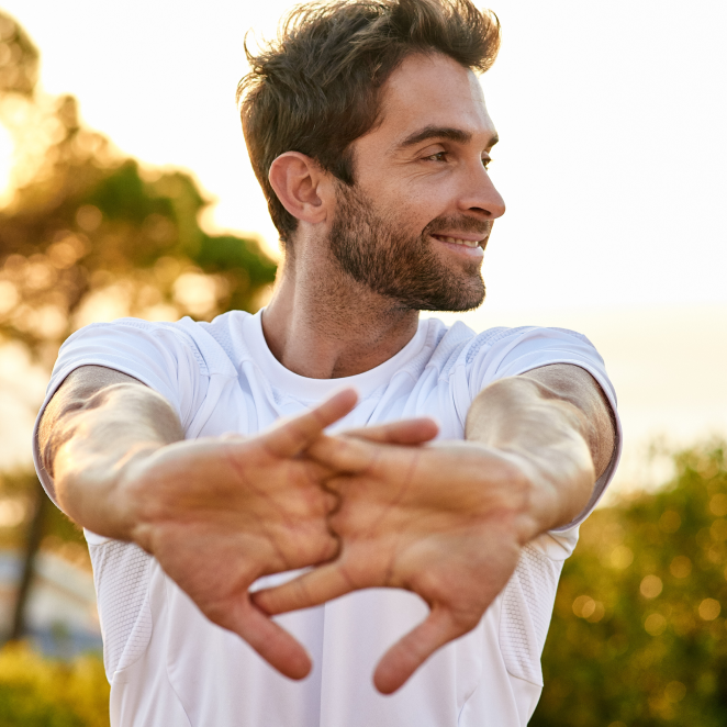 Man in white shirt stretching outdoors with trees in the background.