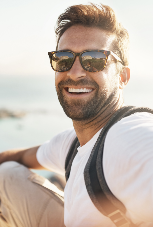 Person smiling in sunglasses, outdoors with sunlight, wearing a white shirt and backpack.