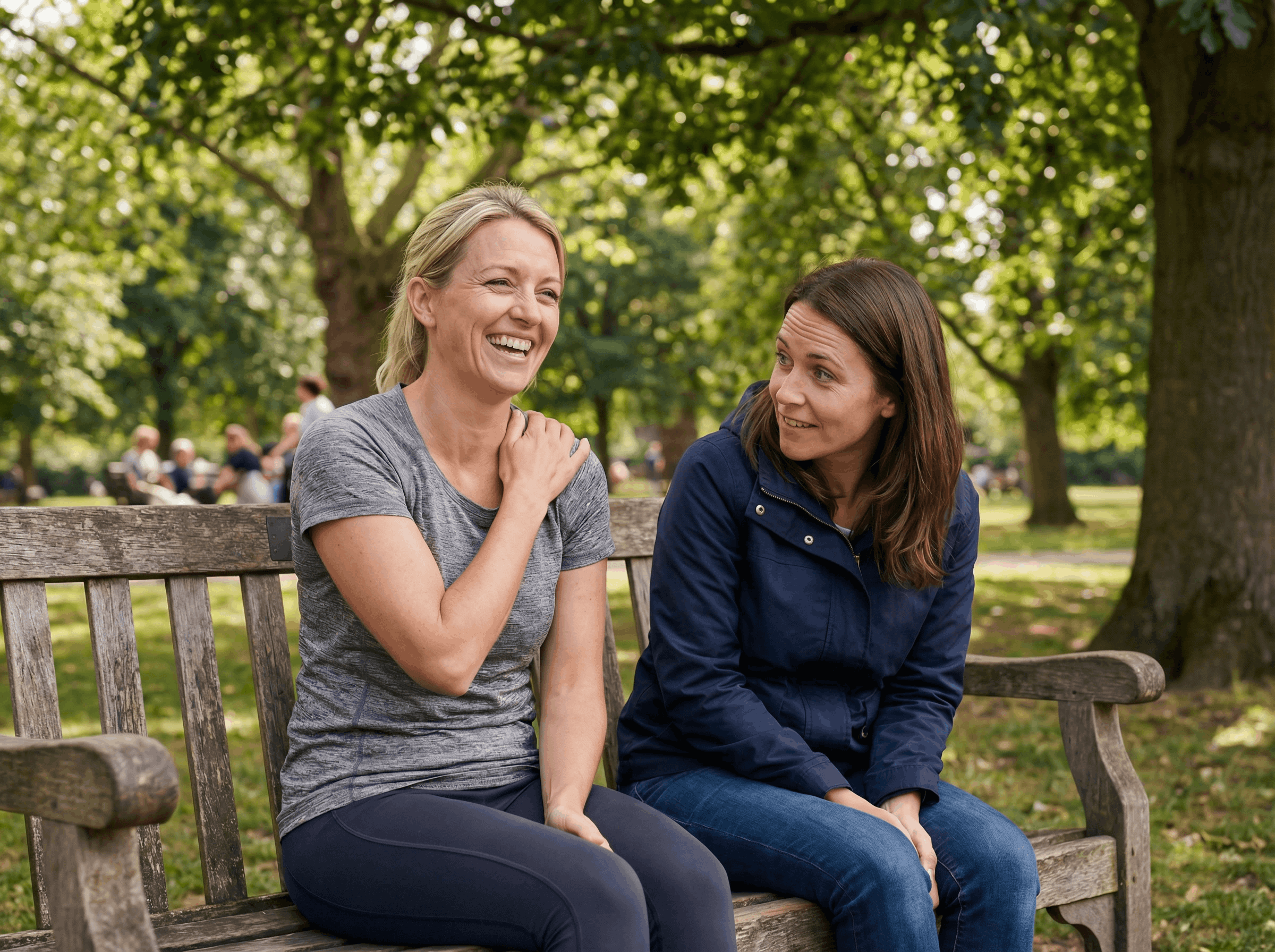 Two women are sitting on a park bench, laughing and talking with one another.