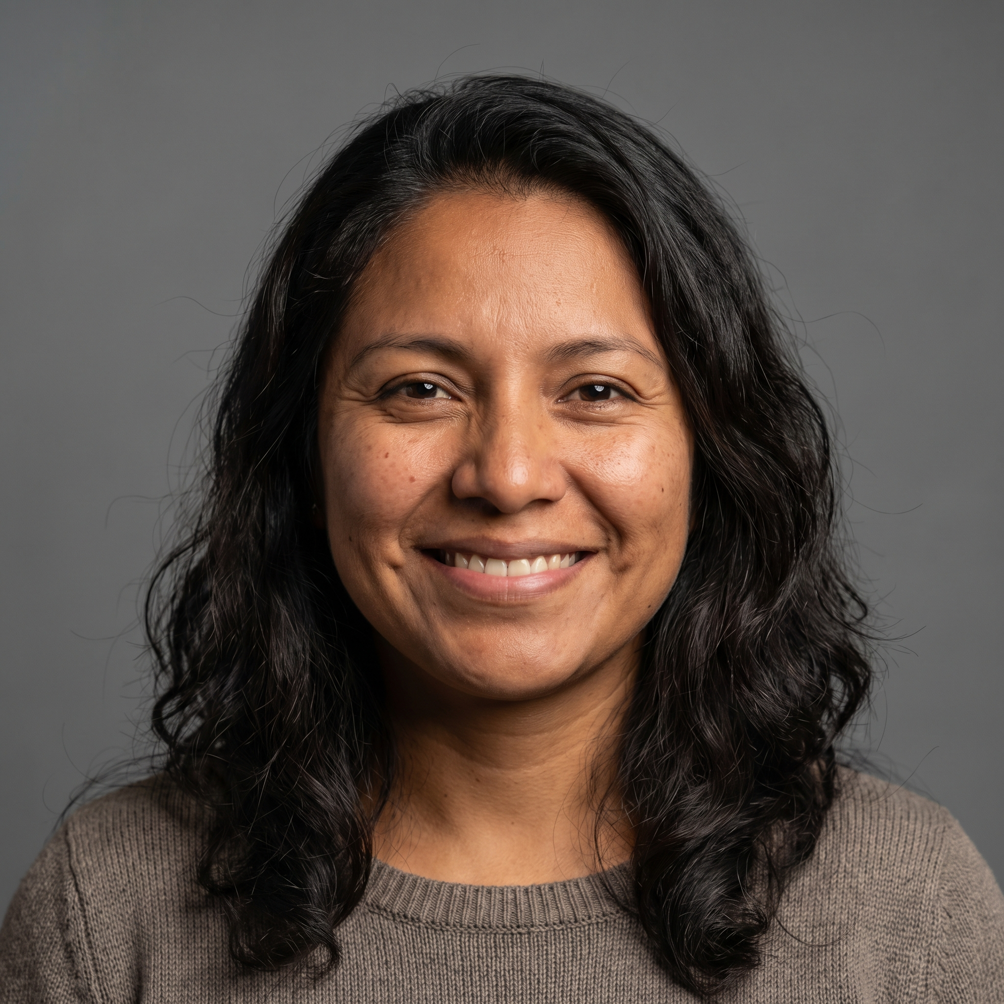 A headshot of a smiling woman with long, dark, wavy hair wearing a brown sweater against a gray background.