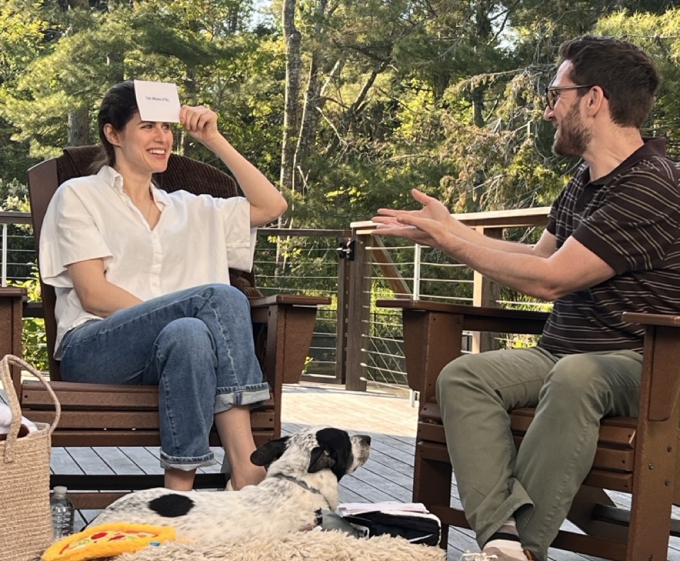 A man and woman sit on a deck with a dog, playing a guessing game with a card.