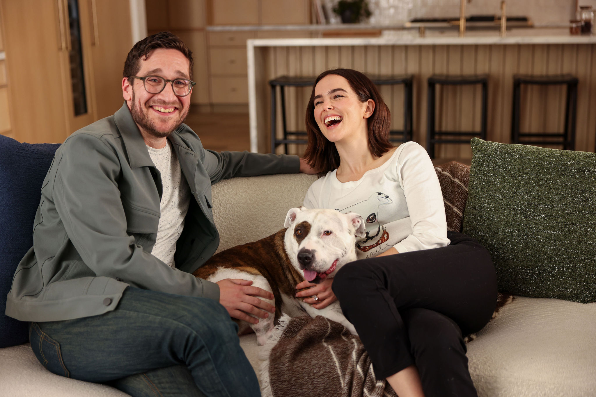 A smiling man and a laughing woman sit on a couch with their dog between them.