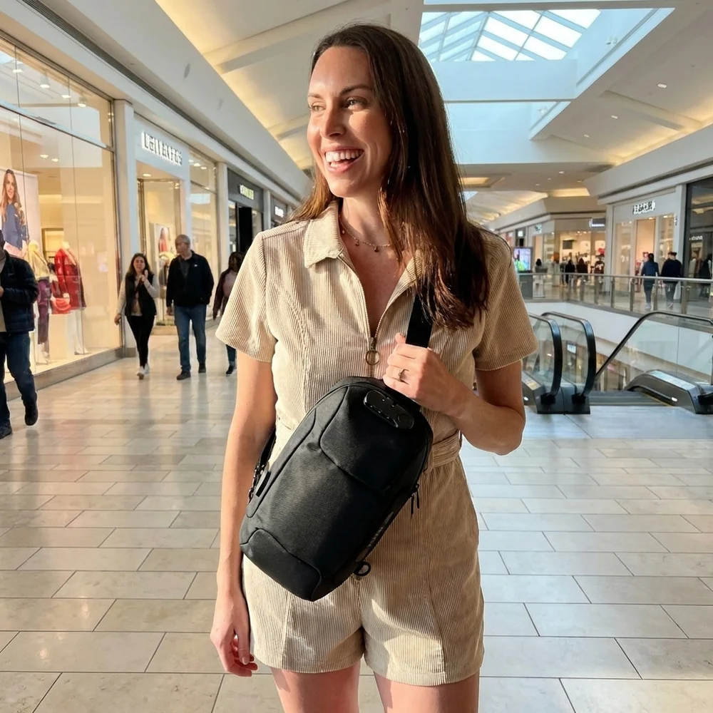 A smiling woman wearing a beige romper and a black sling bag stands inside a shopping mall.