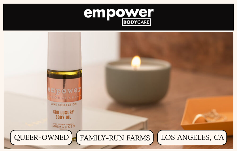 A bottle of Empower Bodycare CBD Luxury Body Oil rests on a surface next to a lit candle.