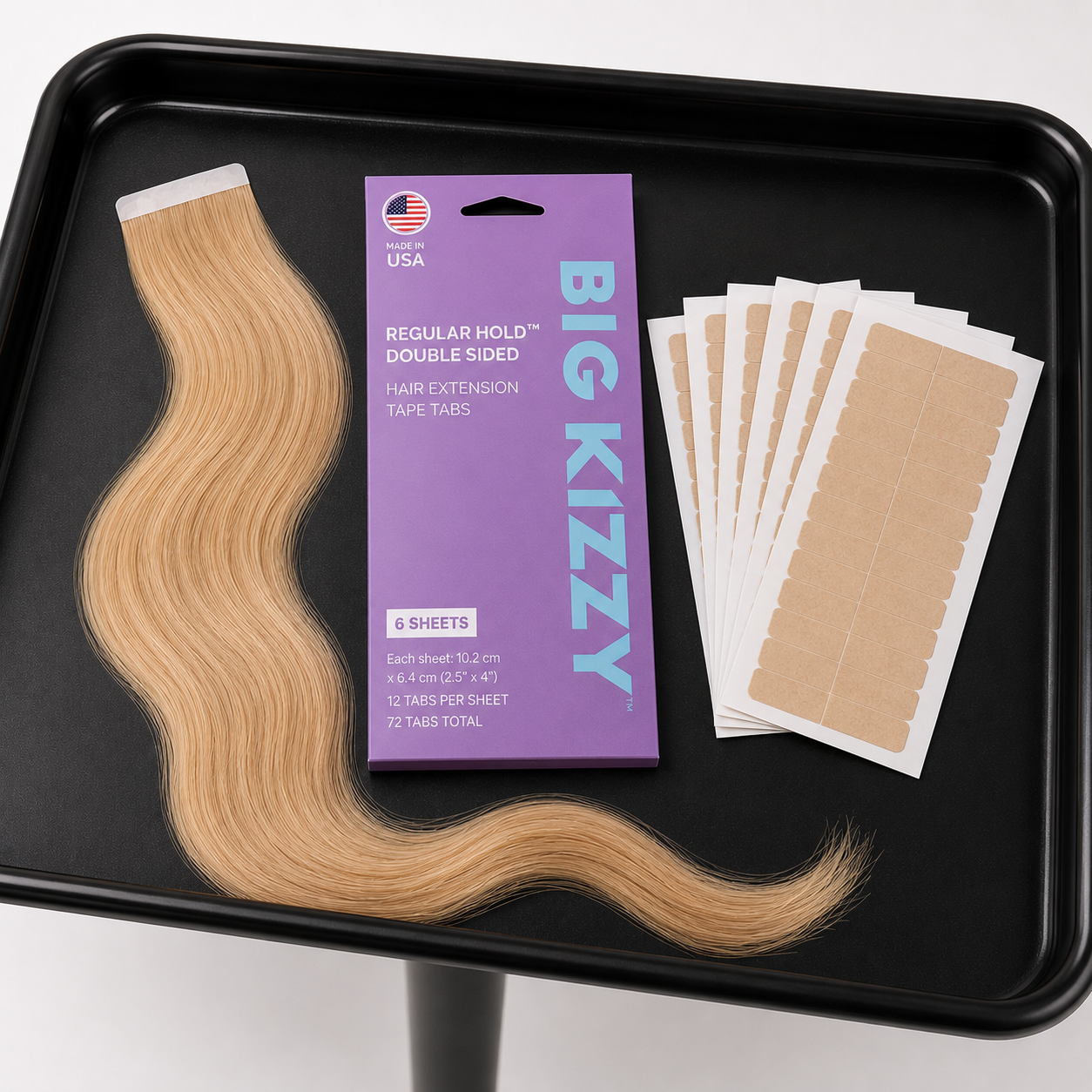 A blonde tape-in hair extension with Big Kizzy packaging and tape tabs on a black tray.