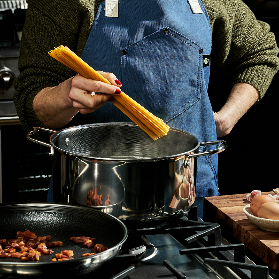 Person in blue apron cooking pasta in a pot on a stove.