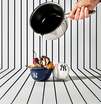 A hand pouring chocolate sauce over ice cream in a baseball-themed bowl with a baseball beside it.