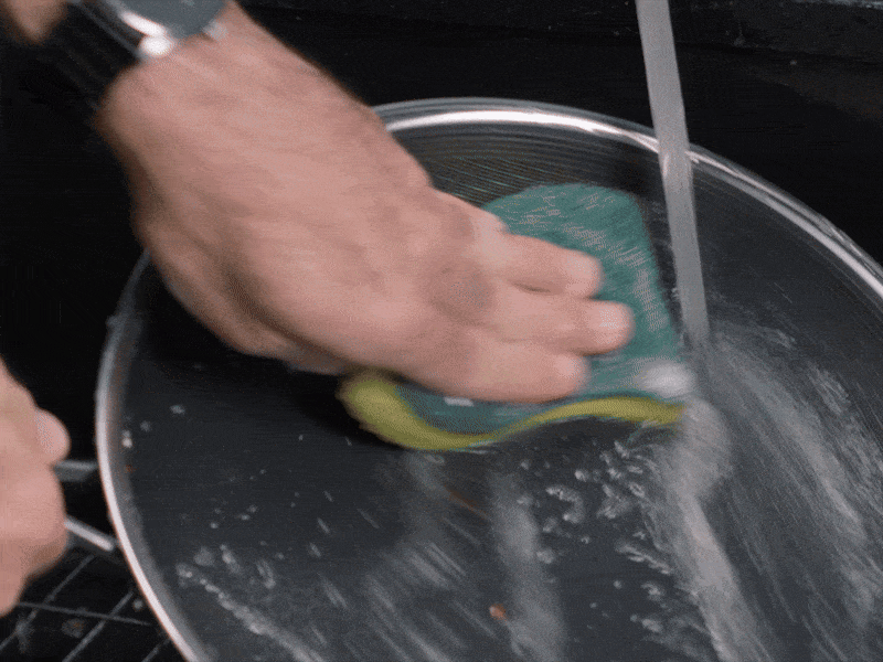 Person scrubbing a pan with a sponge under running water.