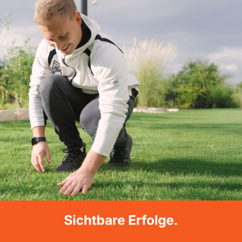 A smiling man in a white and black hoodie kneels on a lush green lawn, touching the grass.